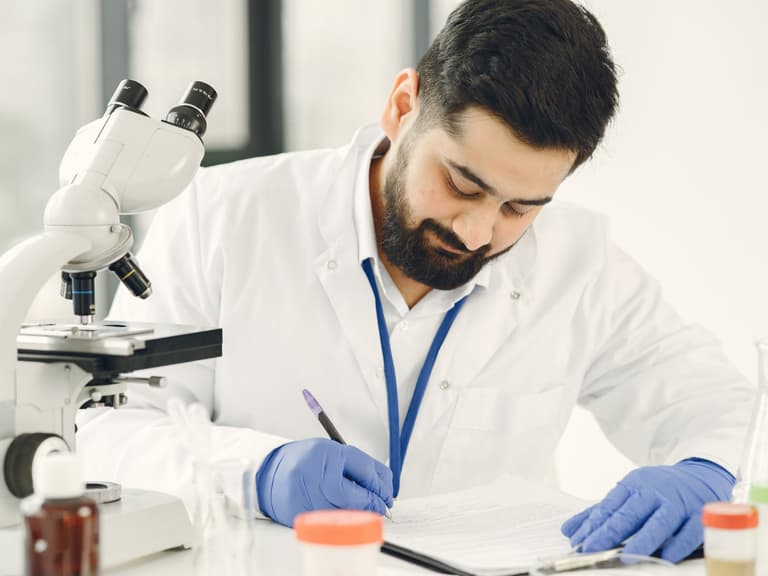 Professionnelle de laboratoire en blouse et gants consignant des résultats près d'un microscope et de tubes d'échantillons